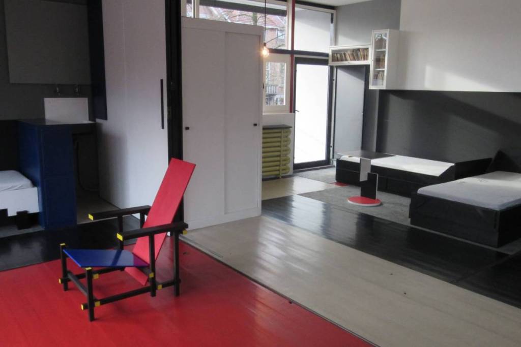 Interieur van het Rietveld Schröderhuis met de iconische Rood-blauwe stoel op een rode vloer, omringd door verschuifbare witte wanden en minimalistische bedden.