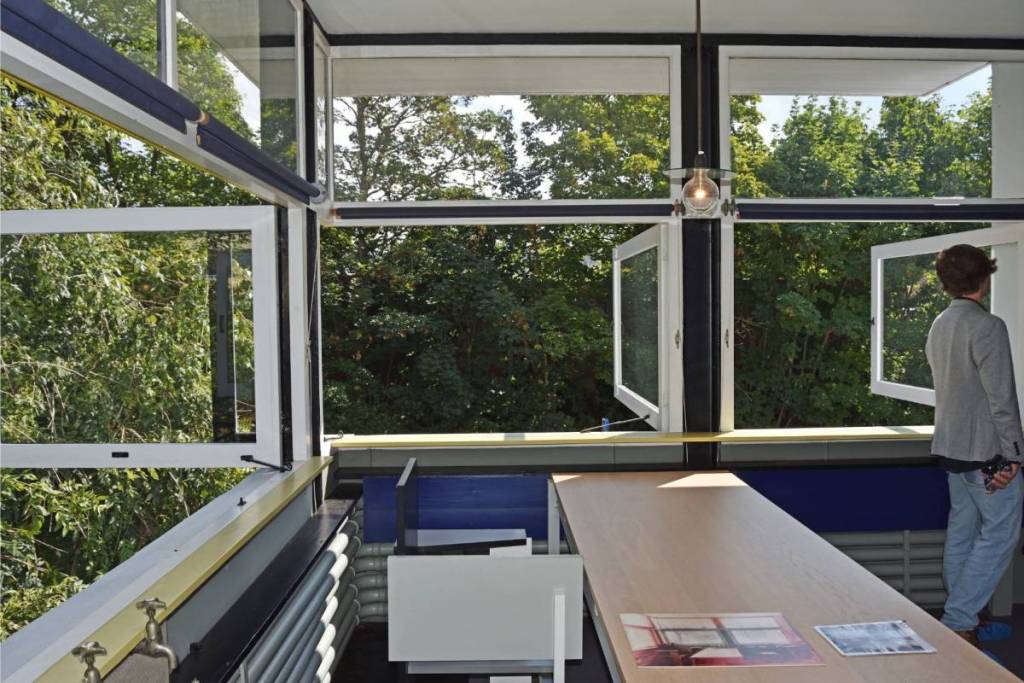 Zicht van binnenuit op het iconische geopende hoekraam van het Rietveld Schröderhuis, waarbij de hoekstijl volledig verdwijnt en uitkijkt op groene bomen. Rechts staat een man die naar buiten kijkt, met op de voorgrond een grote houten tafel en minimalistische meubels.