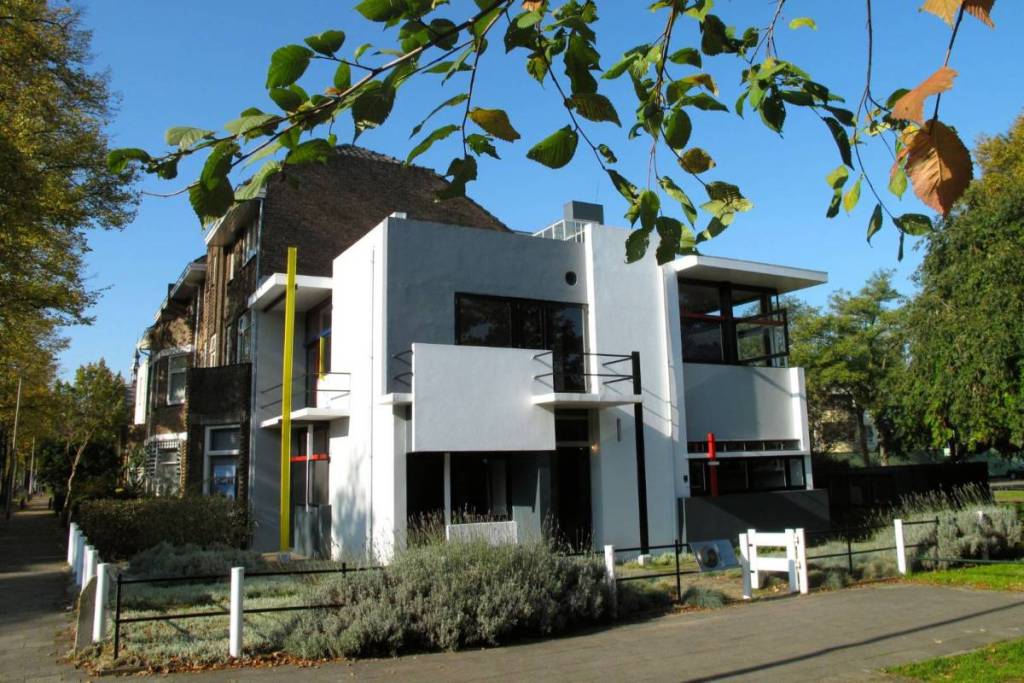 Strakke gevel van het Rietveld Schröderhuis onder een blauwe lucht, deels omlijst door groene boomblaadjes aan de bovenkant en een lavendelstruik op de voorgrond.