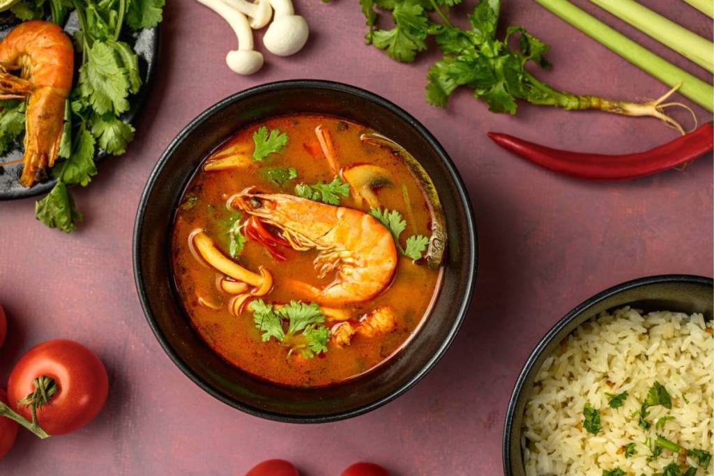 Eine Schüssel würzige thailändische Garnelensuppe (Tom Yum Goong) mit frischem Koriander, Pilzen und einer Beilage aus Reis, fotografiert mit Zutaten wie Zitronengras und Chilischoten.