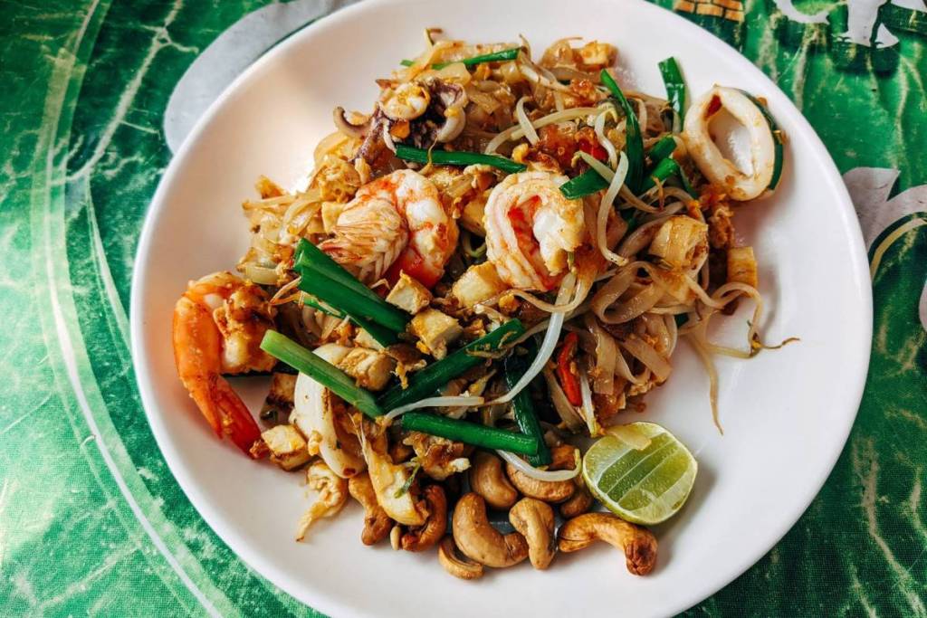 Ein Teller mit authentischem thailändischem Pad Thai, zubereitet mit Garnelen, Tintenfisch, Frühlingszwiebeln und Cashewnüssen, serviert auf einer grünen Tischdecke.