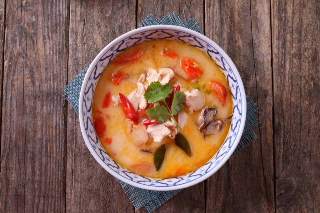 Eine Schüssel cremige thailändische Tom Kha Gai-Hühnersuppe mit Kokosmilch, rotem Chili und frischen Kräutern, serviert auf einem rustikalen Holztisch.