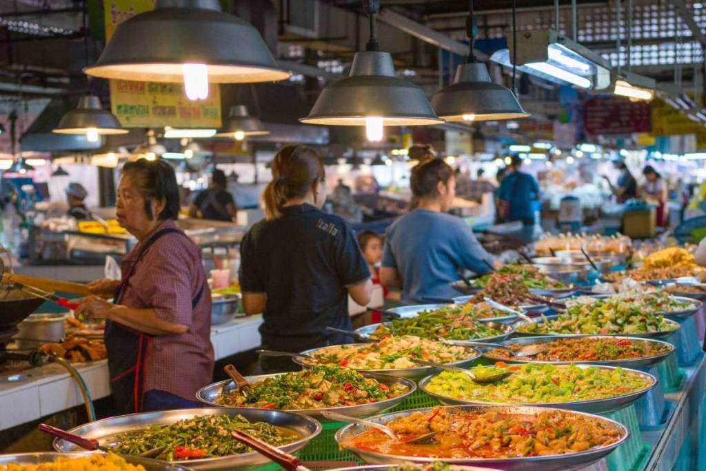 Ein belebter überdachter thailändischer Markt mit großen Metallschalen voller bunter Gerichte und Currys, beleuchtet von Hängelampen, während Mitarbeiter hinter der Theke das Essen zubereiten.