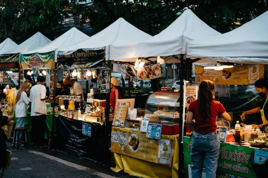 Visitors strolling past illuminated market stalls with white tent canopies and ordering food during a lively evening food festival