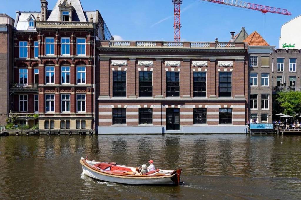 On a sunny day, a small open motorboat with two people on board glides along a wide Amsterdam canal, passing large historic brick buildings and stately facades