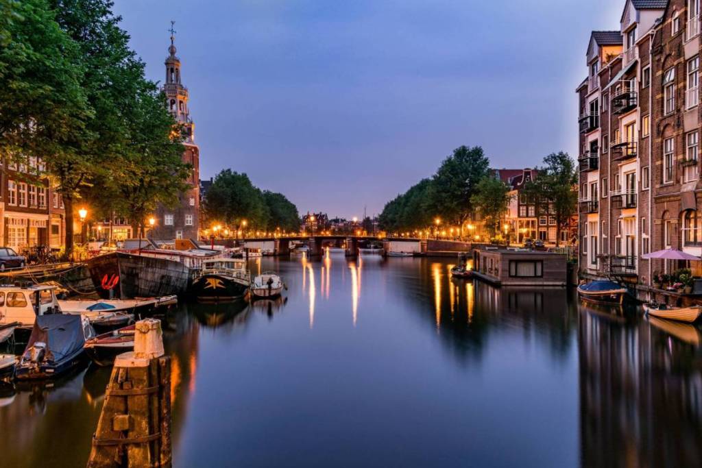 An atmospheric Amsterdam cityscape during the blue hour, with the illuminated historic Montelbaanstoren on the left and traditional canal-side houses on the right along the mirror-smooth waters of the Oudeschans