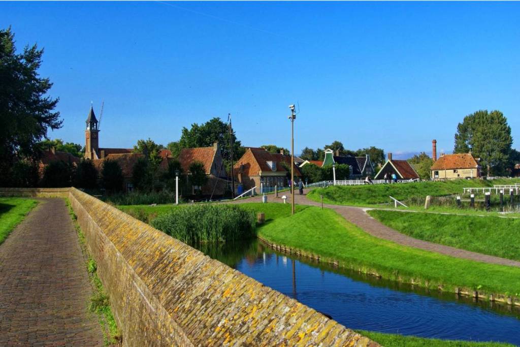 Eine steinerne Stadtmauer schlängelt sich entlang eines Kanals und über Wiesen in Richtung der historischen Häuser und des Kirchturms von Enkhuizen.