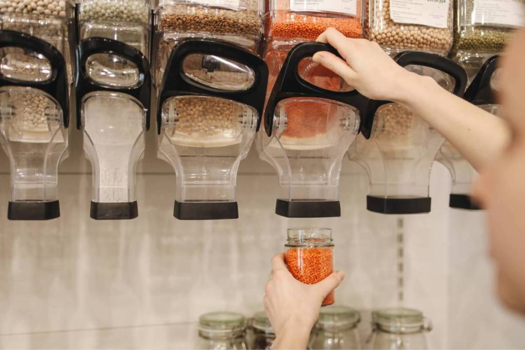 Jemand füllt rote Linsen aus einem durchsichtigen Spender in ein Glas in einem verpackungsfreien Bioladen.