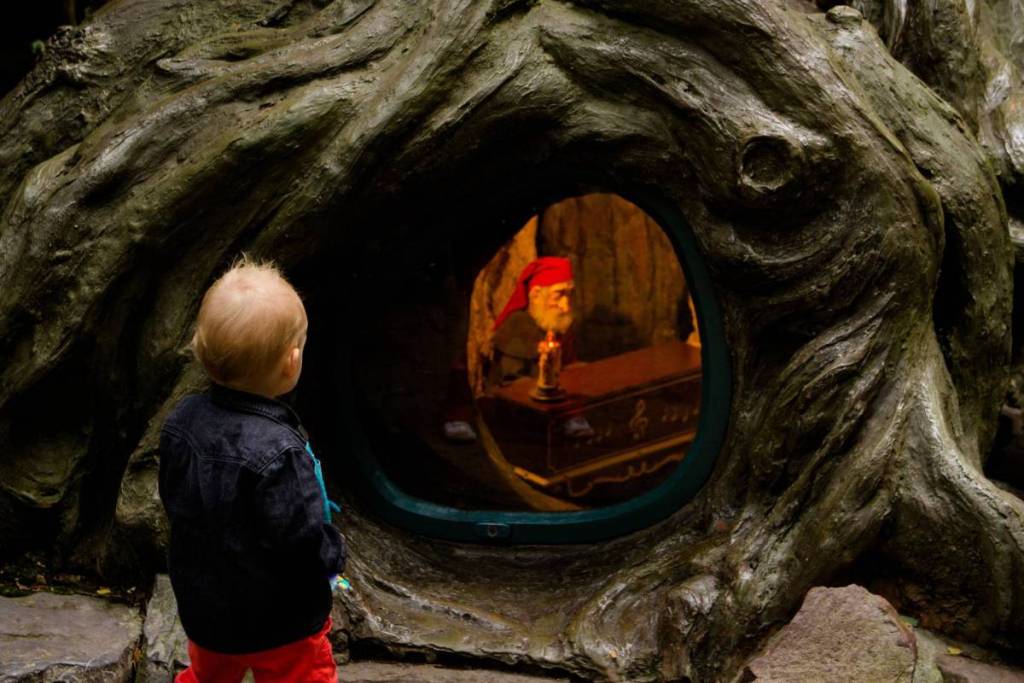 A toddler looks curiously at a gnome in a hollow tree trunk in the Gnome Village of the Efteling Fairy Tale Forest