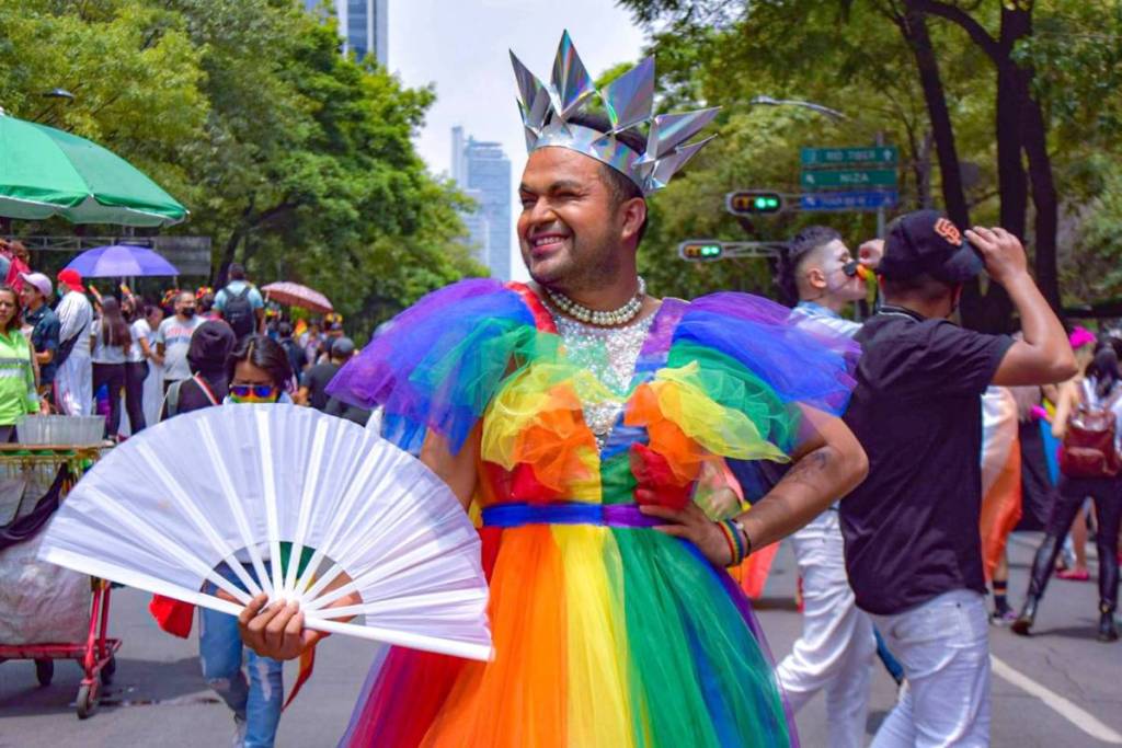 Un participante sonriente con un colorido vestido arcoíris, una corona plateada y un gran abanico blanco durante la Pride Walk.