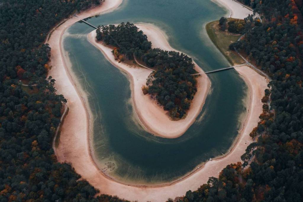 Una foto aerea dell’esclusivo lago Henschotermeer a forma di ferro di cavallo con la sua isola centrale, circondato da fitti boschi e vaste spiagge sabbiose.