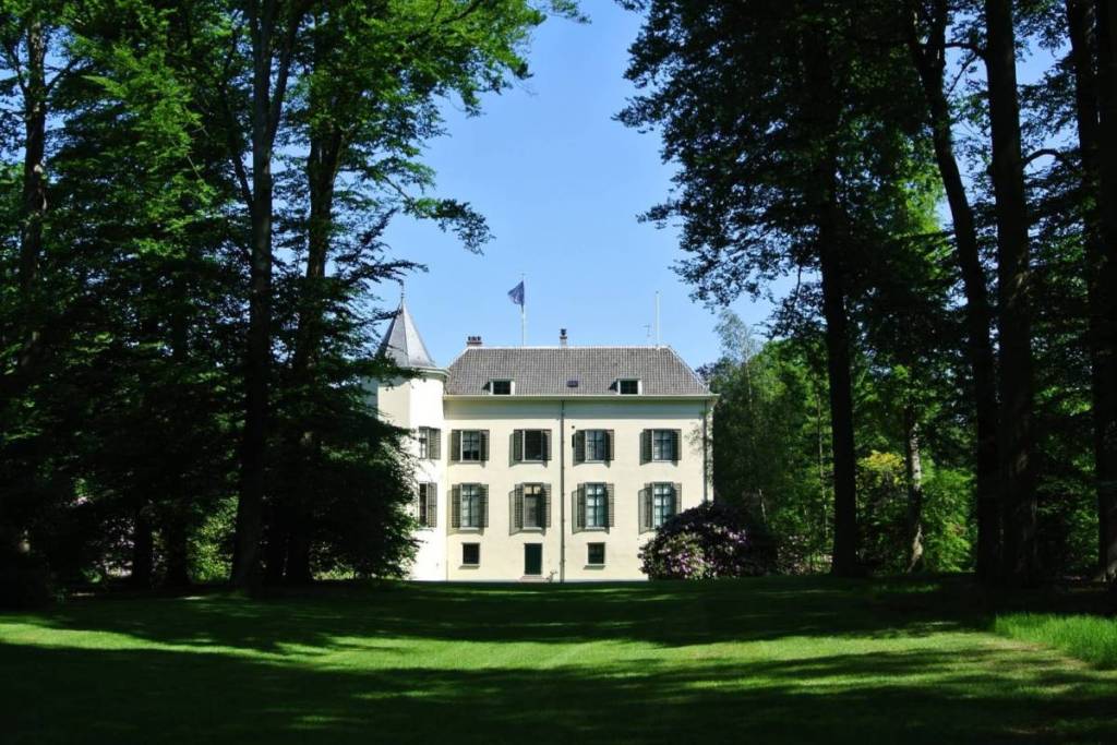 Vue de profil du manoir blanc Huis Doorn, entouré de grands arbres et d’une vaste pelouse verte sur le domaine.
