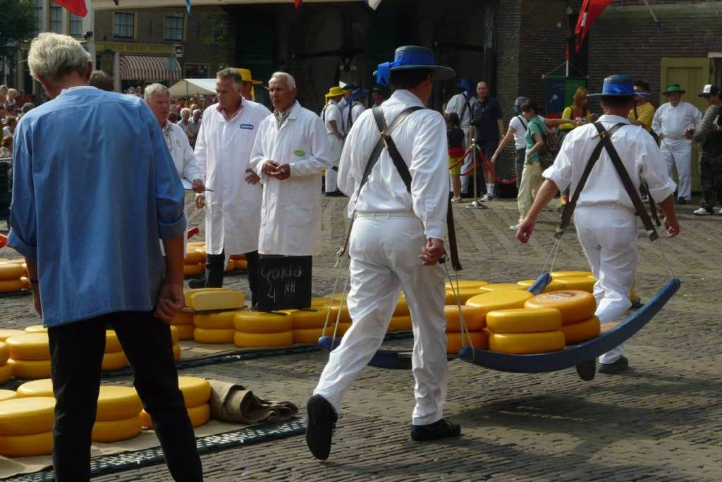Ein Übersichtsfoto vom Alkmaarer Käsemarkt auf dem Waagplein, wo zwei Mitglieder der Käseträgergilde mit blauen Hutbändern einen prall gefüllten blauen Korb tragen. Im Vordergrund liegen Stapel von Gouda-Käsen, und ein Mann in einem blauen Hemd schaut zu.