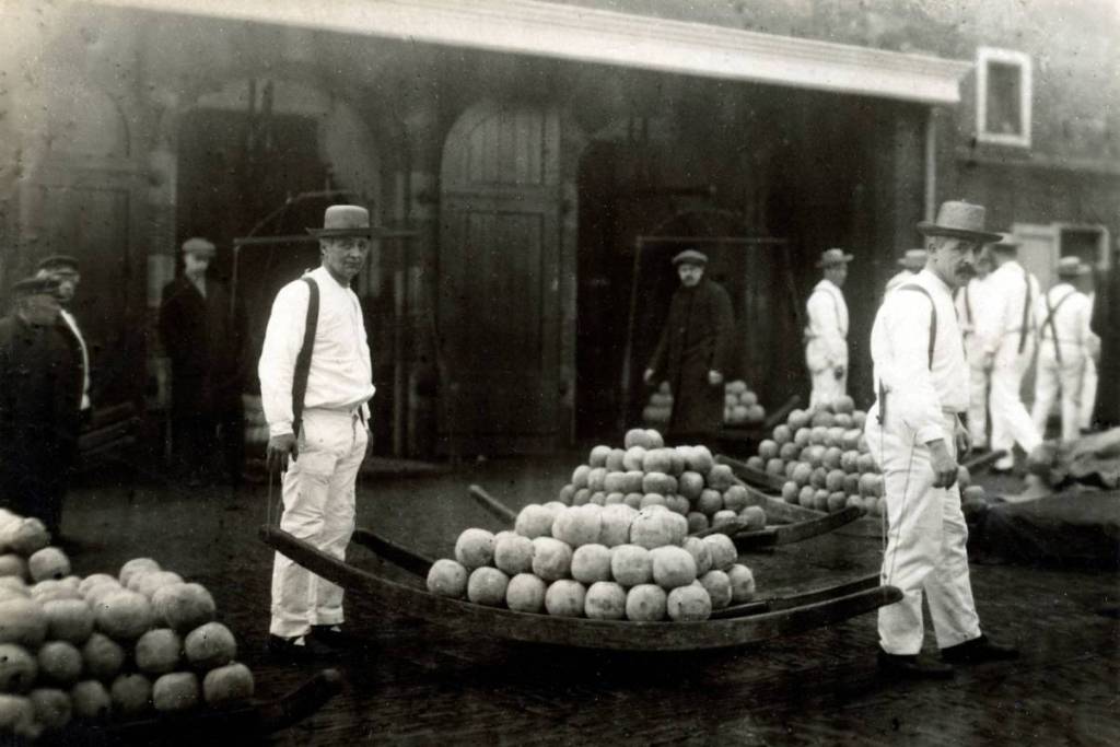Ein historisches Schwarz-Weiß-Foto vom Alkmaarer Käsemarkt, das zwei Mitglieder der Käseträger-Gilde in traditioneller weißer Kleidung und mit Hüten zeigt, wie sie vor dem Wiegehaus neben Käsestücken und Stapeln runder Käse posieren.