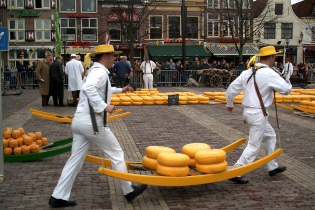 Zwei Mitglieder der Käseträgergilde mit gelben Hutbändern tragen im Laufschritt eine gelbe Trage mit Gouda-Käsen über den gepflasterten Waagplein in Alkmaar, vor dem Hintergrund historischer Gebäude.