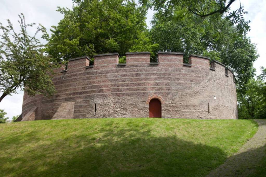 Detalle de la muralla medieval de ladrillo de la fortaleza de Leiden, con almenas, aspilleras y una puerta de acceso roja.