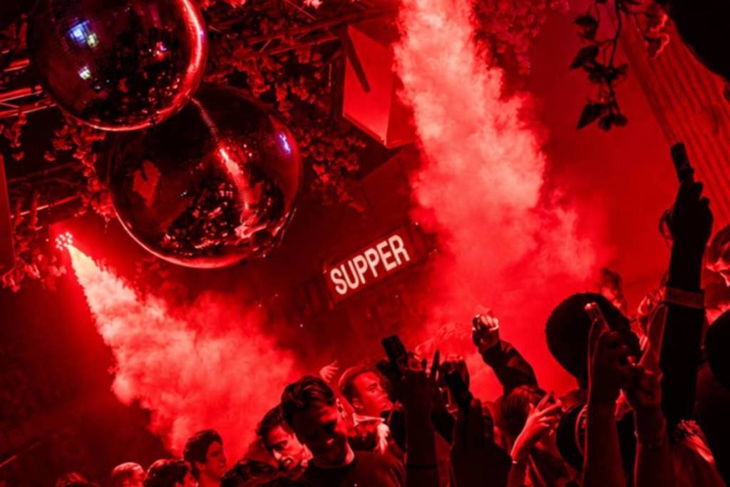 Público de fiesta frente a un letrero de neón iluminado de «SUPPER» con efectos de humo rojo.
