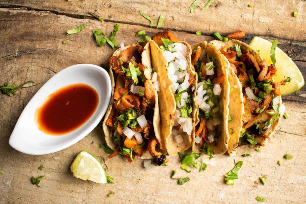 Three fresh Mexican tacos with marinated meat, finely chopped onion, cilantro, and a small bowl of red salsa on a wooden platter.