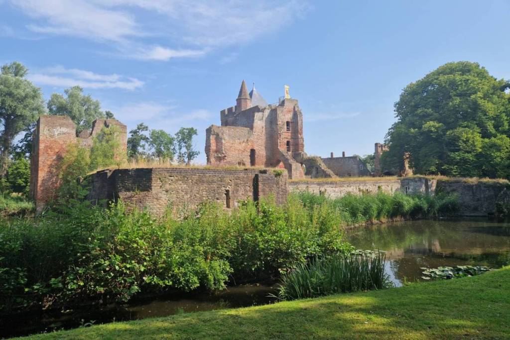 Una vista soleggiata sulle rovine storiche in mattoni del Castello di Brederode, circondato da un fossato, da una vegetazione rigogliosa e da alberi sotto un cielo azzurro.