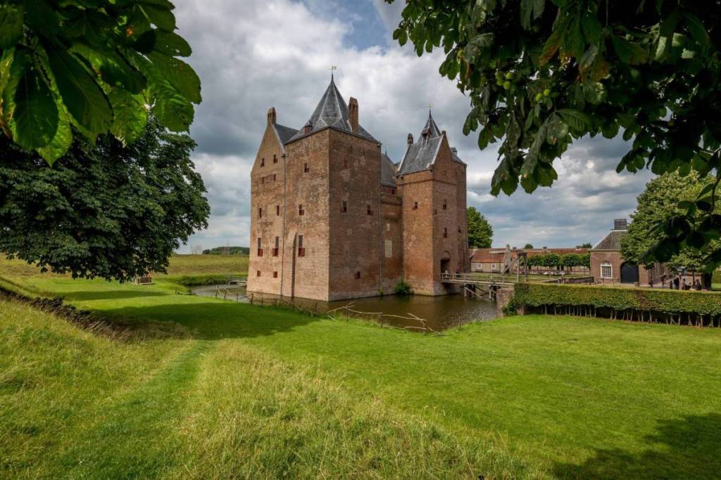El castillo medieval de Loevestein, con sus torres puntiagudas y sus contraventanas rojas, rodeado por un foso con un puente de madera y un césped verde.