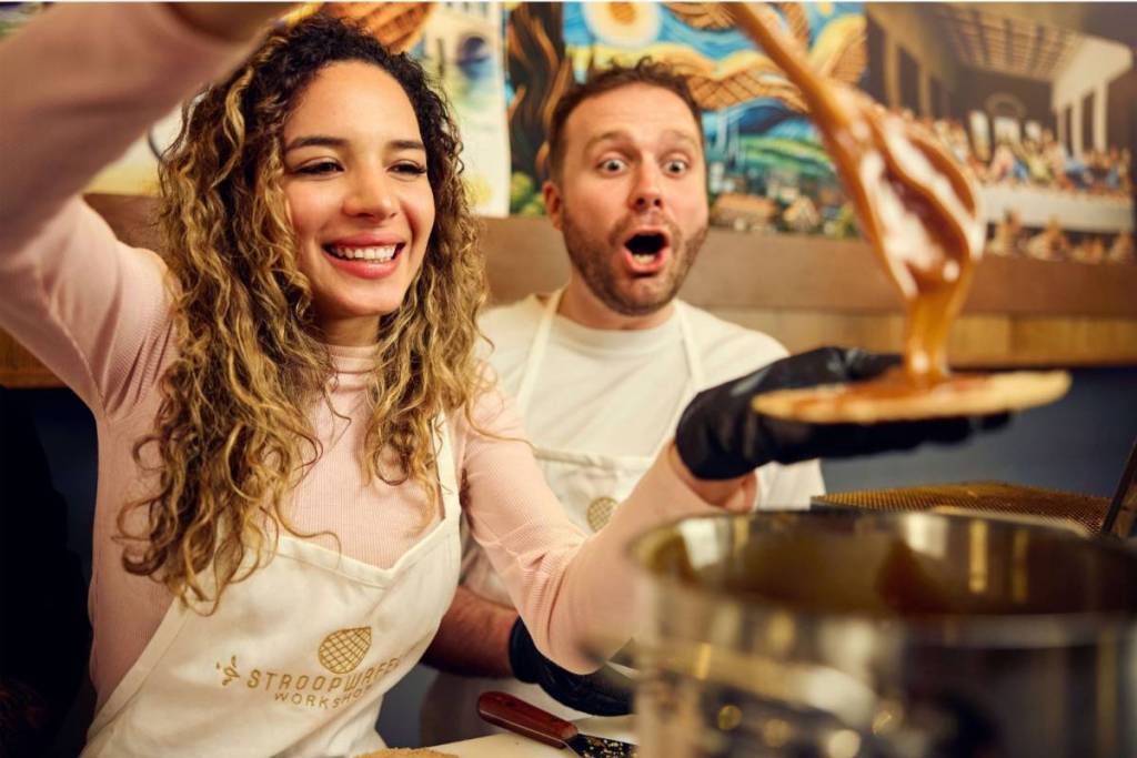 Een lachende vrouw die warme karamel op een verse stroopwafel giet, terwijl een man in een schort verbaasd toekijkt tijdens een workshop.