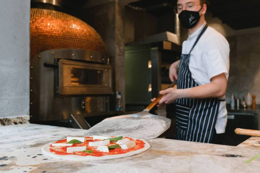 Een pizzabakker met mondkapje gebruikt een schep om een ongebakken pizza Margherita naar de pizza-oven te verplaatsen.