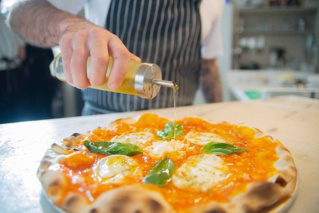 Een pizzabakker die olijfolie giet over een versgebakken pizza met mozzarella en basilicum.