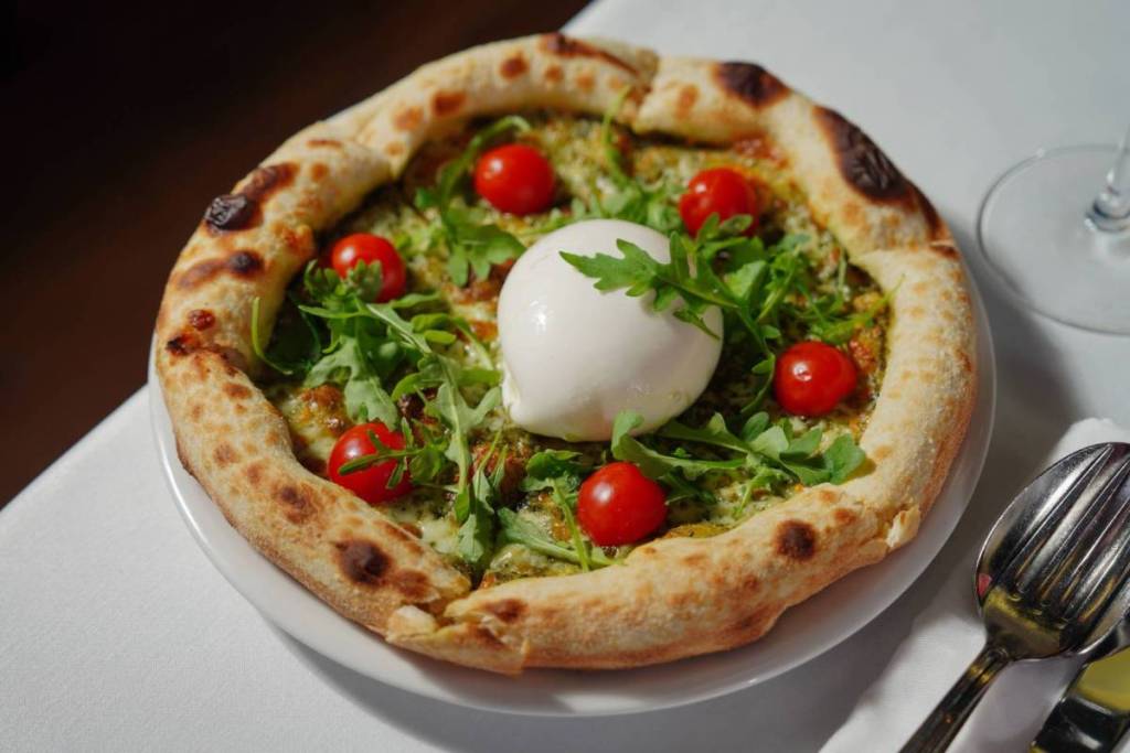 Verse Neapolitanse pizza op een bord, belegd met pesto, rucola, kerstomaten en een grote bol burrata in het midden.