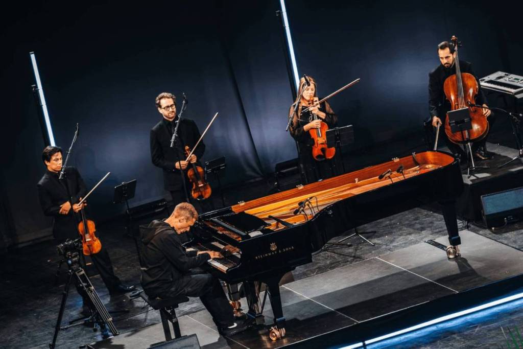 Una toma desde arriba de Armin van Buuren tocando un piano de cola Steinway, rodeado por el cuarteto de cuerda Hydra Quartet en un escenario iluminado.