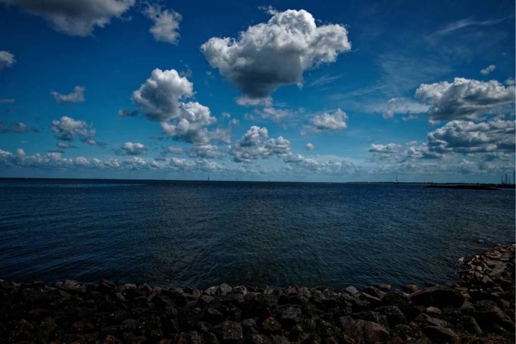Vue sur les eaux bleu foncé de l’IJsselmeer depuis une digue en pierre, sous un ciel bleu parsemé de nuages blancs.