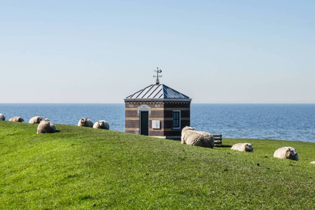 Des moutons couchés sur une digue verdoyante près d’une petite maison d’observation historique en briques au bord de l’IJsselmeer.