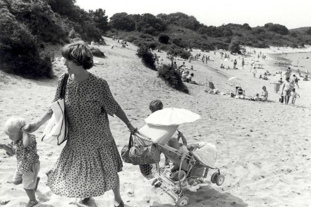 Foto en blanco y negro de una mujer que camina con un niño pequeño y un cochecito por la playa de arena del lago de dunas «'t Wed», con gente disfrutando del tiempo libre al fondo.