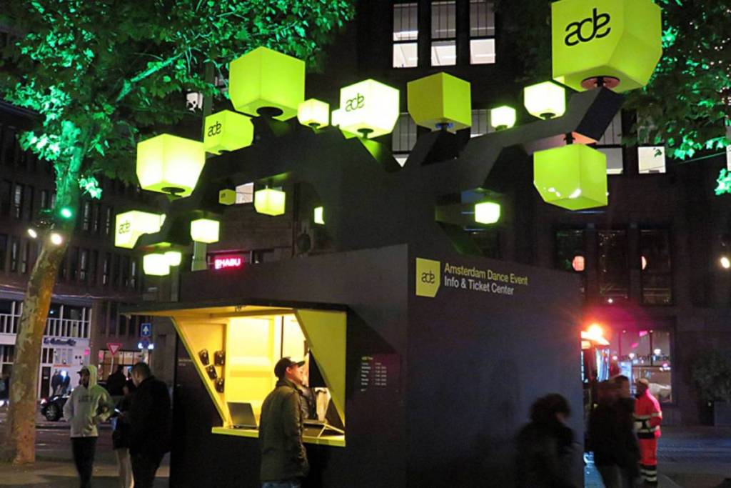 Het verlichte Amsterdam Dance Event Info & Ticket Center op straat in het donker, gedecoreerd met opvallende gele ADE-kubuslampen.