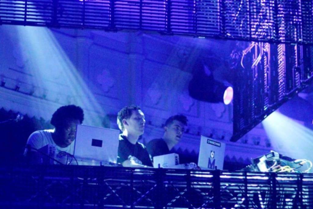 Drie artiesten achter hun laptops en apparatuur op een verhoogd podium onder blauwe spotlights.