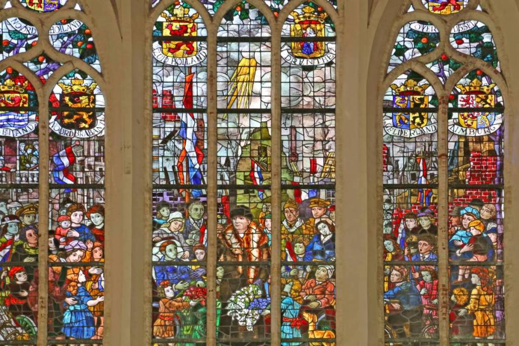 Colorida vidriera de la Oude Kerk con detalladas escenas históricas, banderas neerlandesas y escudos provinciales.