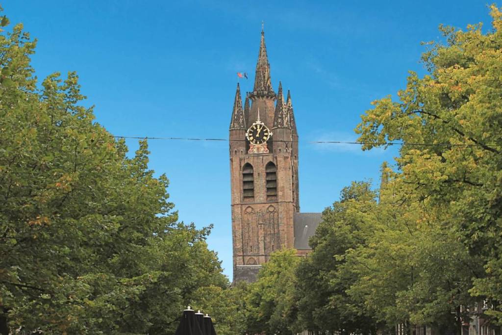 La torre inclinada de la Oude Kerk («Scheve Jan») en Delft, que se alza por encima de una hilera de árboles verdes bajo un cielo azul claro.
