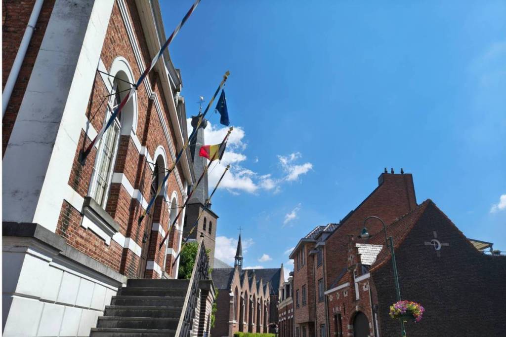 Vue de rue à Baarle avec des bâtiments historiques en briques rouges, des drapeaux belges et de l'UE hissés et un clocher d'église en arrière-plan.