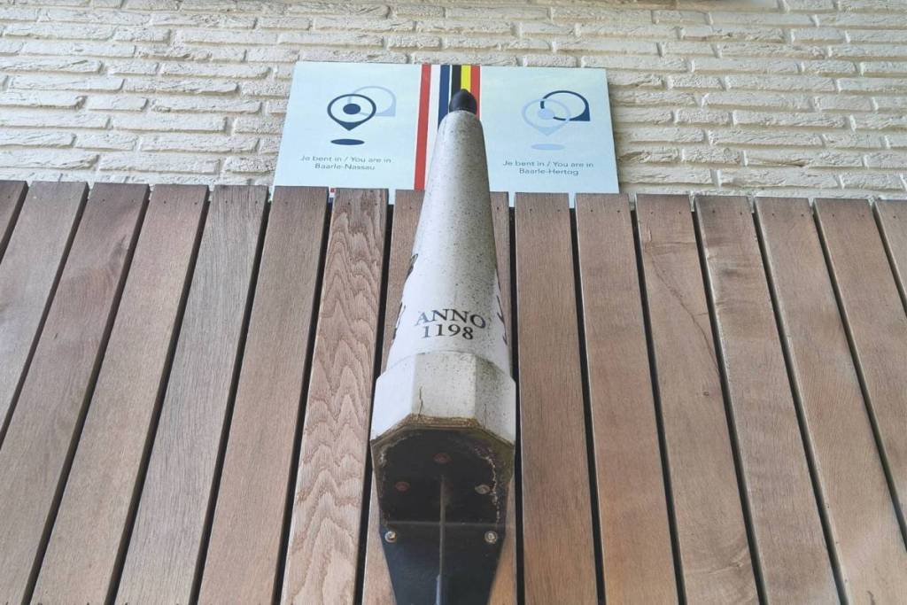 Réplique d’une borne frontalière portant l’inscription « Anno 1198 », fixée sur un panneau en bois sous un panneau d’information indiquant la frontière entre Baarle-Nassau et Baarle-Hertog.