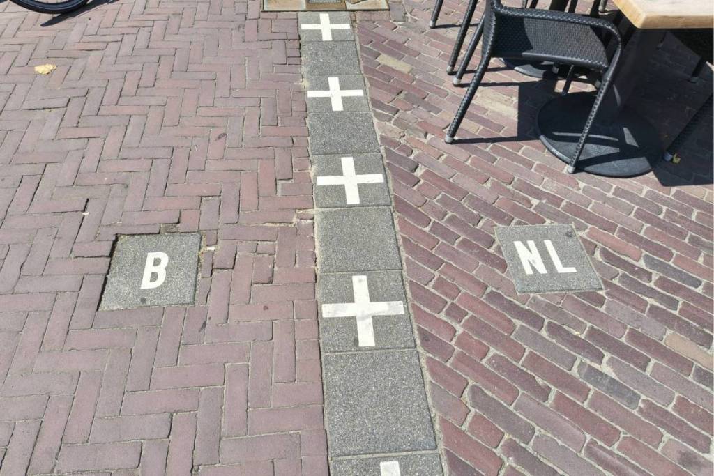 Une ligne de frontière clairement marquée sur le pavage avec des dalles « B » (Belgique) et « NL » (Pays-Bas) près d’une terrasse.