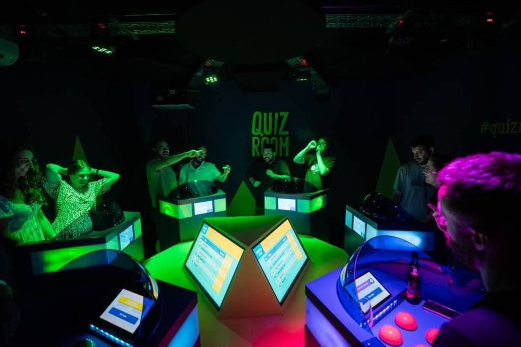 Un animado grupo de amigos juega en la interactiva Quiz Room de Middelburg, iluminada por luces de neón de color verde intenso y rodeada de cabinas de concurso con pantallas.