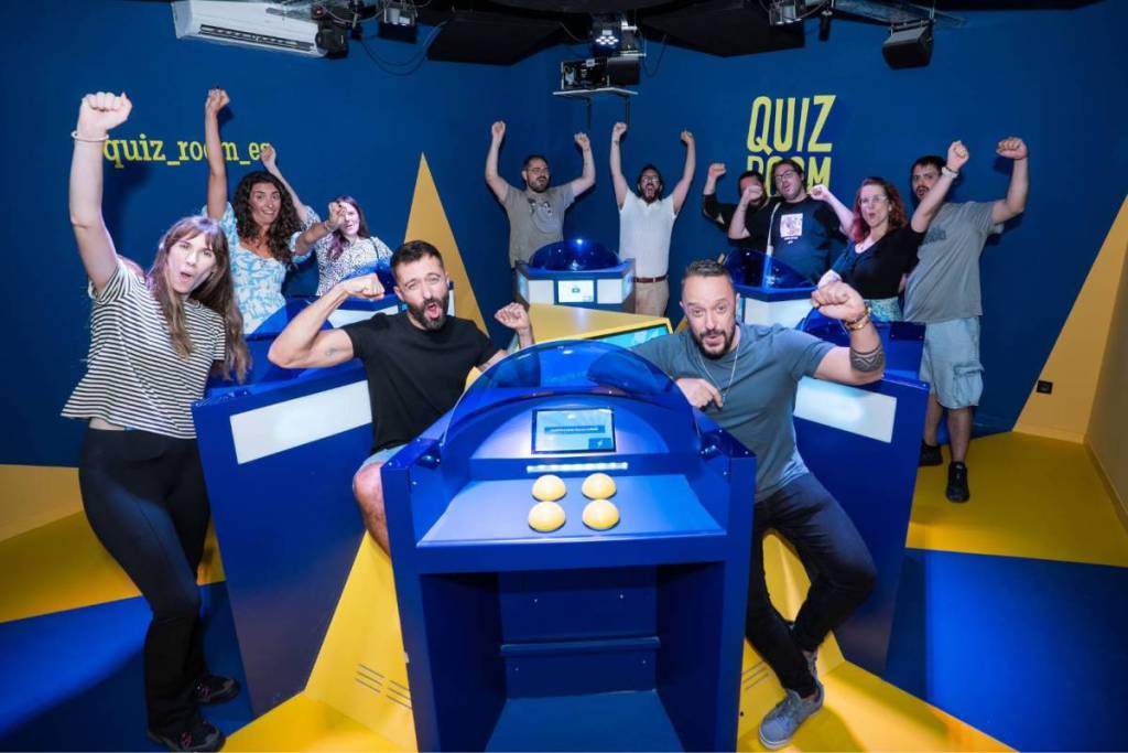 Once personas súper entusiasmadas celebran su victoria en el Quiz Room de Middelburg con los puños en alto, de pie alrededor de una estación de quiz.