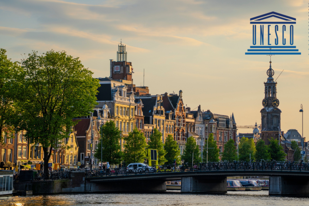 Klassische Amsterdamer Grachtenkulisse mit dem Munttoren im Hintergrund und dem offenen blauen UNESCO-Weltkulturerbe-Logo in der rechten oberen Ecke.