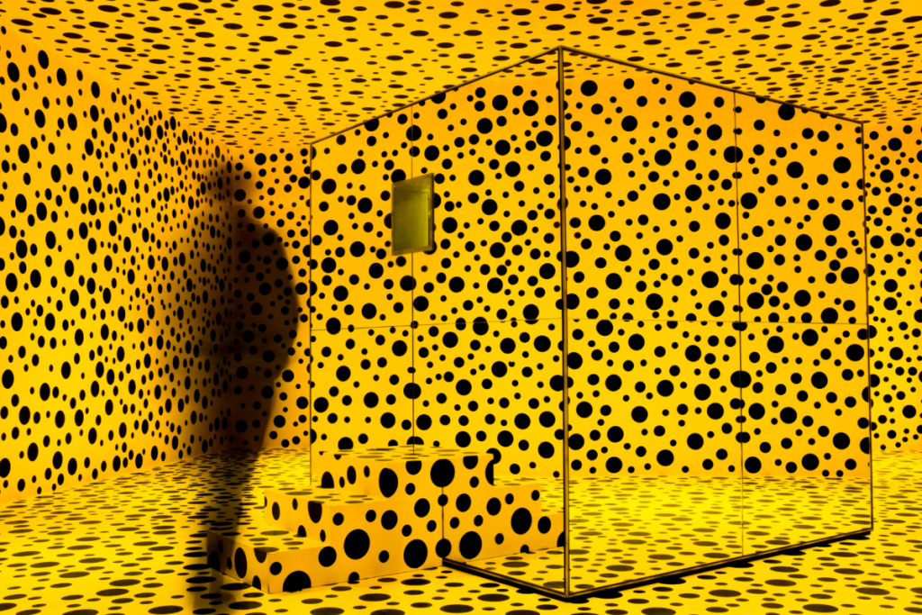 Exterior of a mirrored cube in a room with yellow-and-black polka-dotted walls and floor.