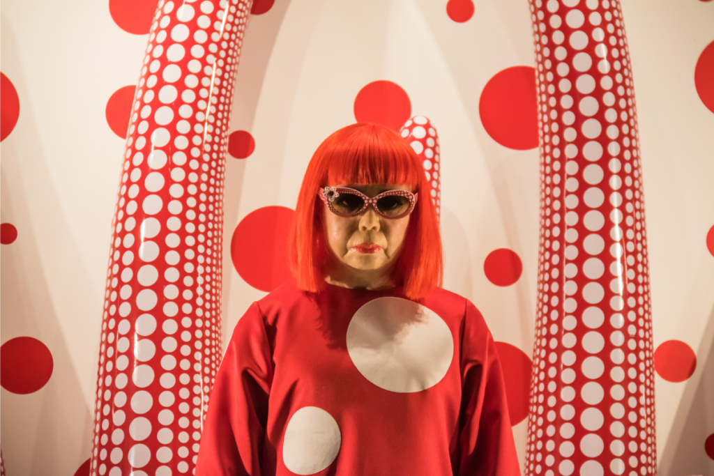 Porträt der Künstlerin Yayoi Kusama in einem roten Polka-Dot-Kleid, mit Sonnenbrille und roten Haaren, stehend neben weiß-rot gemusterten Skulpturen.