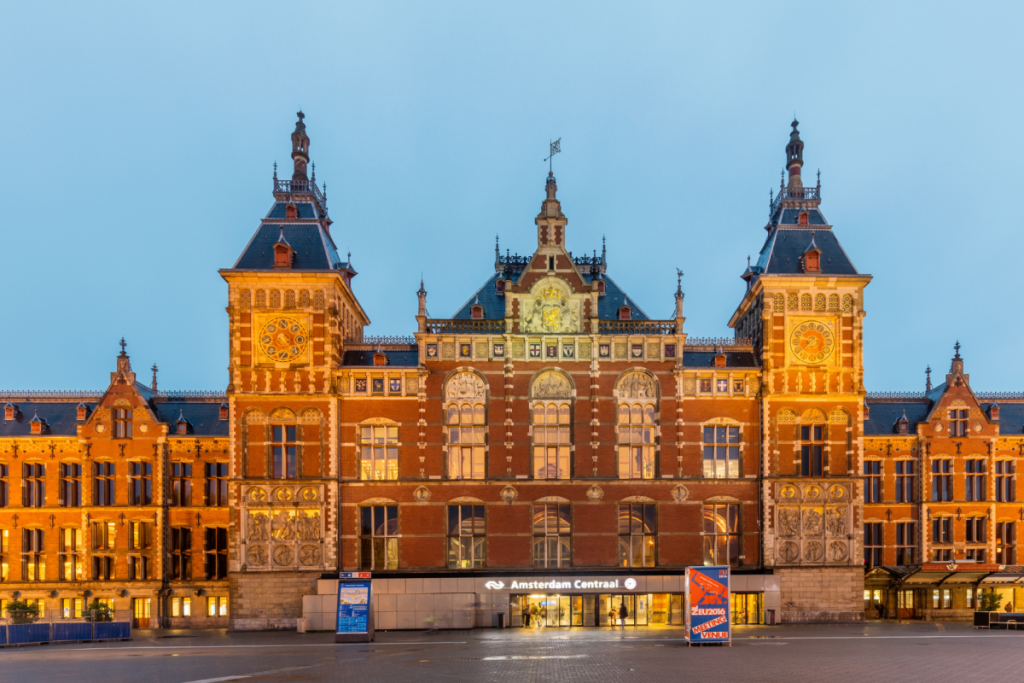 Vorderansicht der neugotischen Fassade des Amsterdamer Hauptbahnhofs mit beleuchteten Türmen, entworfen von Pierre Cuypers.