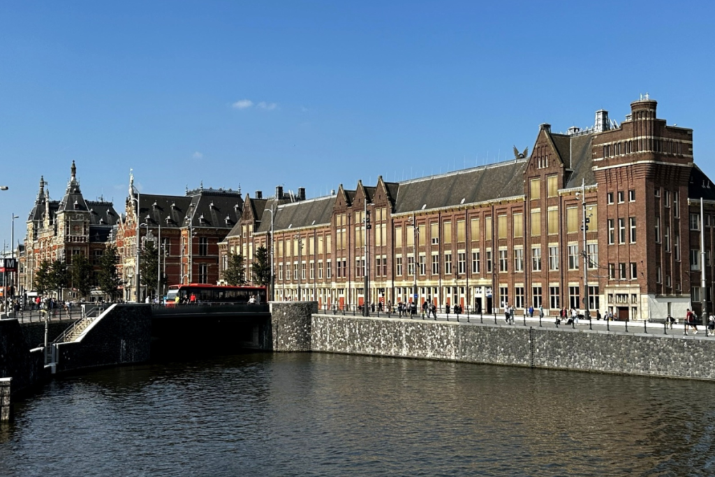 Seitenansicht des Bahnhofs Amsterdam Centraal am Wasser, mit Blick auf das Backstein-Bahnhofsgebäude und den Kai.