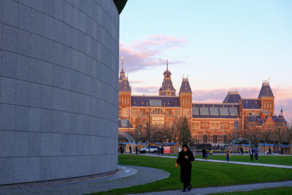 Das ikonische Rijksmuseum in Amsterdam bei Sonnenuntergang, gesehen vom grünen Museumplein aus, mit einem Hauch moderner Architektur im Vordergrund.