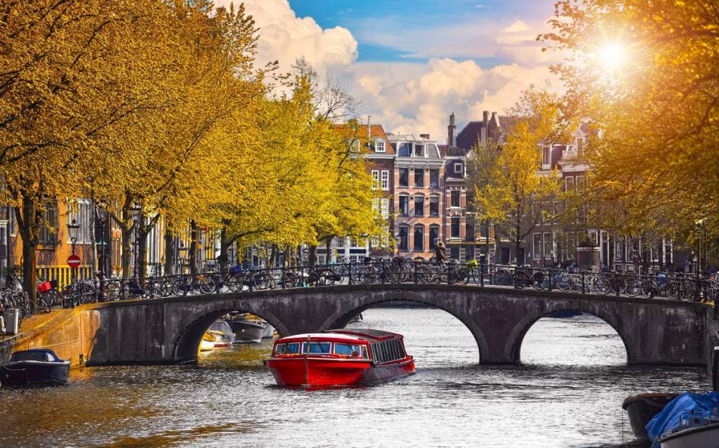 Eine typische Amsterdamer Grachtenkulisse im Herbst mit einem roten Wassertaxi unter einer mit Fahrrädern übersäten Bogenbrücke, umgeben von goldfarbenen Bäumen und traditionellen Grachtenhäusern im Sonnenlicht.