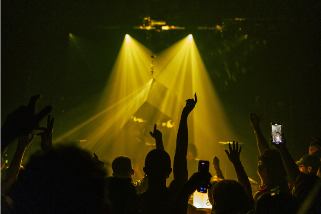 Siluetas del público de fiesta con las manos en alto junto a un escenario iluminado por focos amarillos durante el Amsterdam Dance Event 2026.