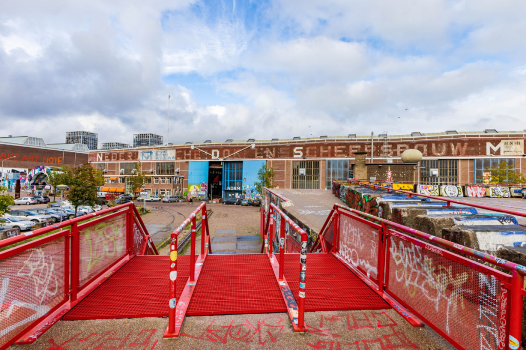 Blick auf die ikonische Außenfassade der NDSM-Halle von der roten Fußgängerbrücke auf dem NDSM-Werftgelände aus.